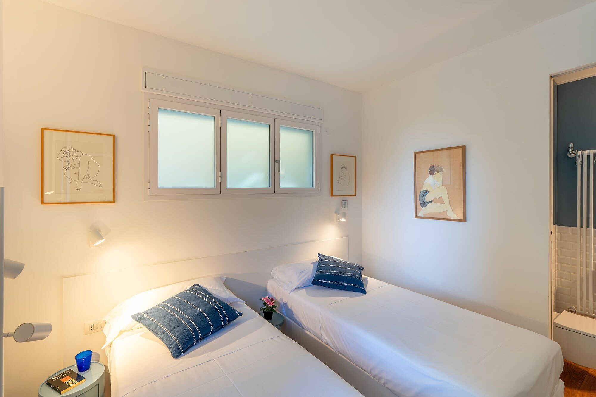 Una camera d'albergo con due letti, una finestra con vetro satinato, un'opera d'arte appesa al muro, un comodino con una lampada e un termosifone.