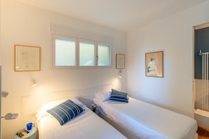 Una camera d'albergo con due letti, una finestra con vetro satinato, un'opera d'arte appesa al muro, un comodino con una lampada e un termosifone.