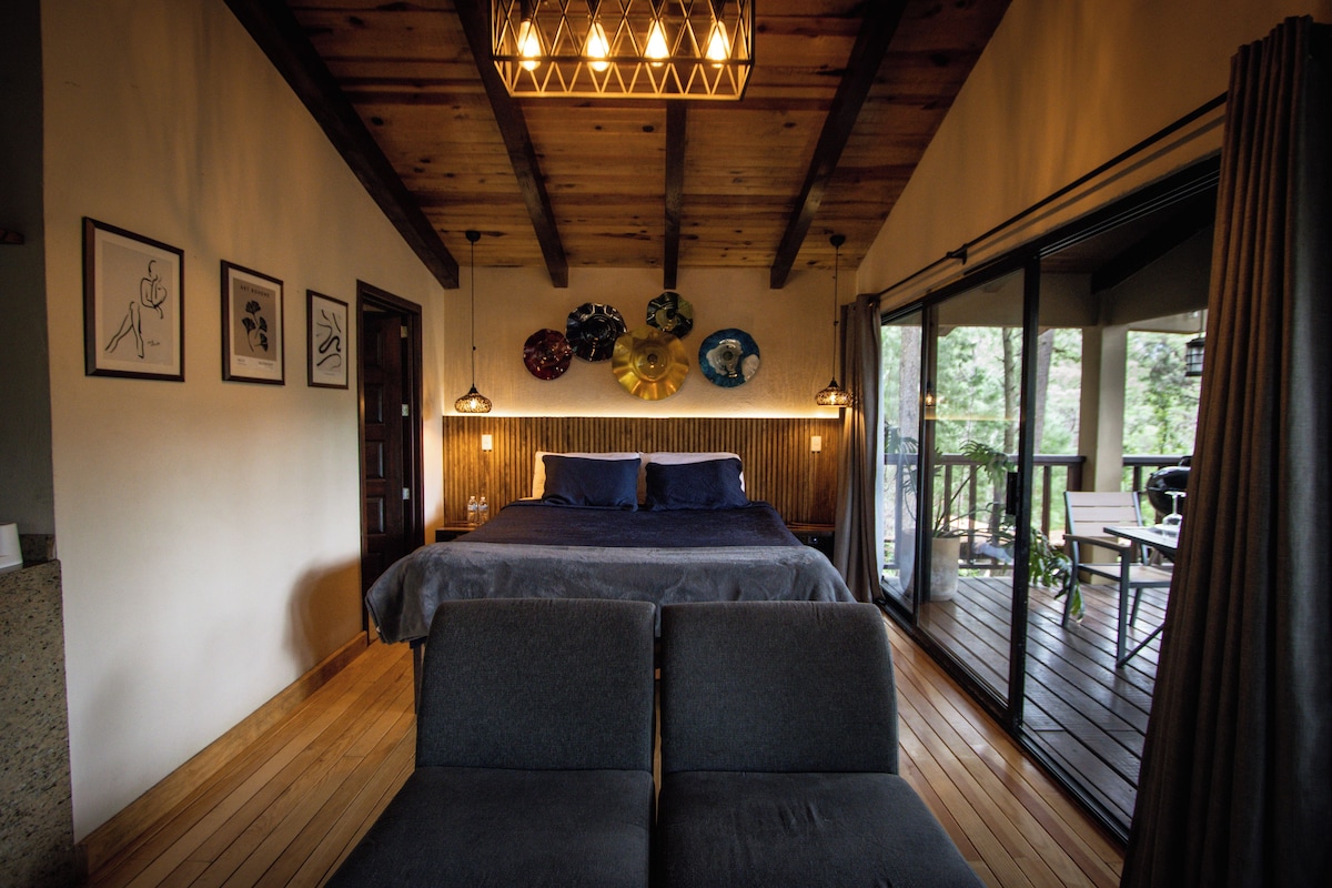 A bedroom with a large bed, two armchairs, a wooden ceiling, and a view of the outdoors through a sliding door.