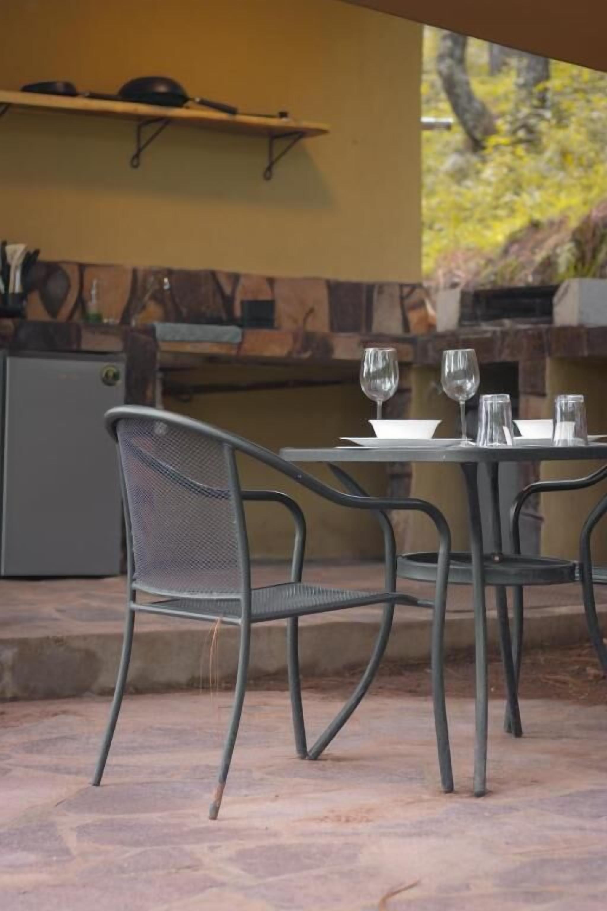 Un área de comedor al aire libre con sillas de metal y una mesa, suelo de piedra y una cocina al fondo.