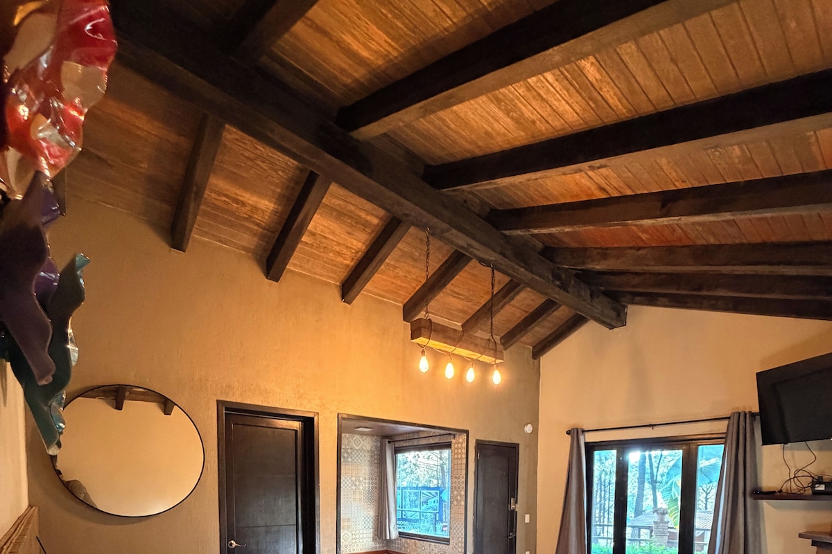 A bedroom with a large bed, a wooden ceiling, a round mirror, and a window with a view of the outdoors.
