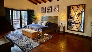 Un dormitorio con una cama grande, techo de madera, un ventanal amplio y un espejo geométrico.