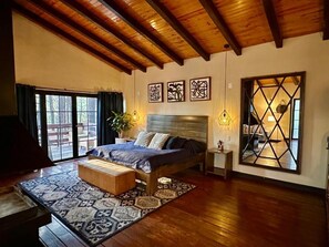 A bedroom with a large bed, a wooden ceiling, a large window, and a geometric mirror.