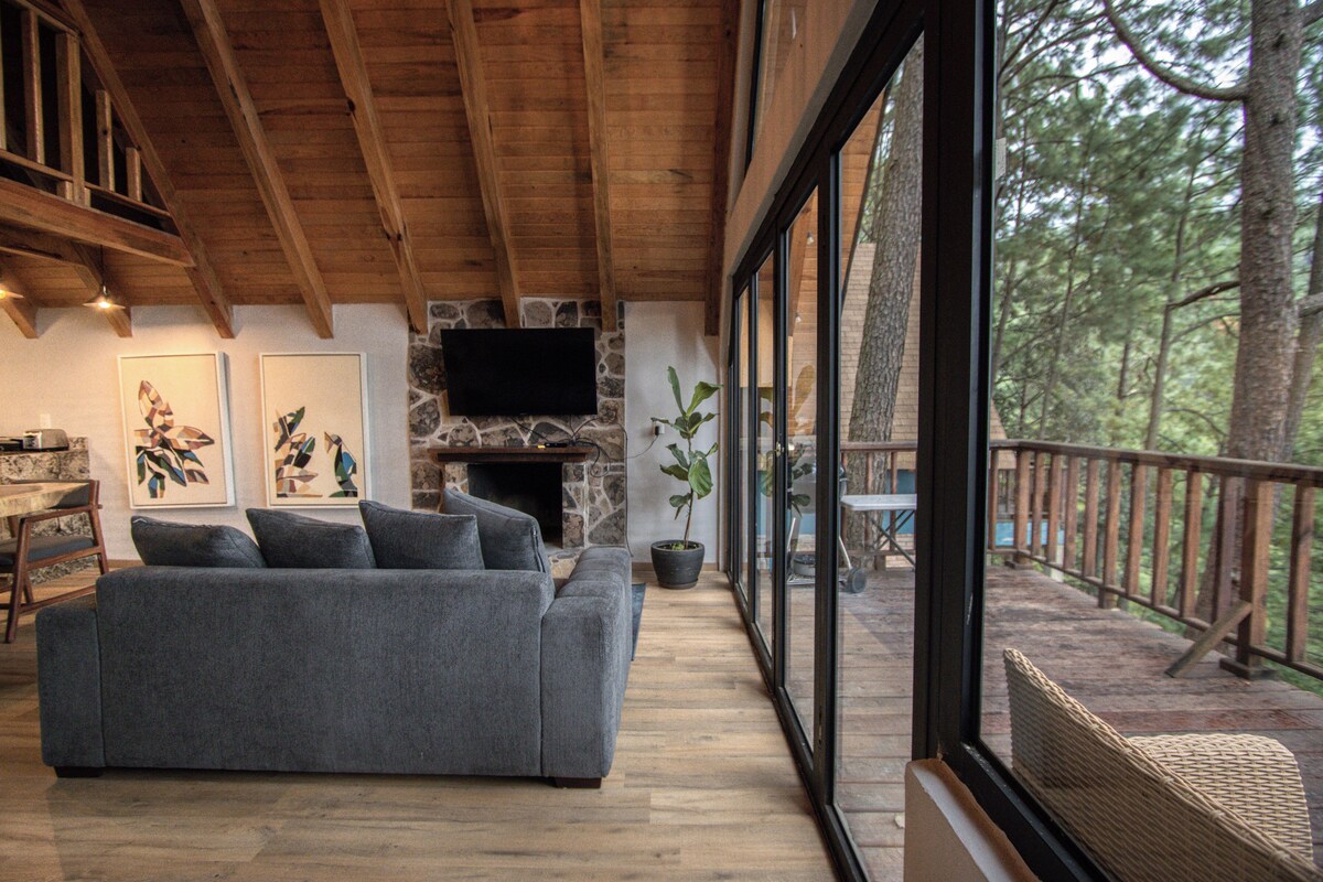 A cozy living room with a gray sofa, a stone fireplace, and a large window overlooking a forest.