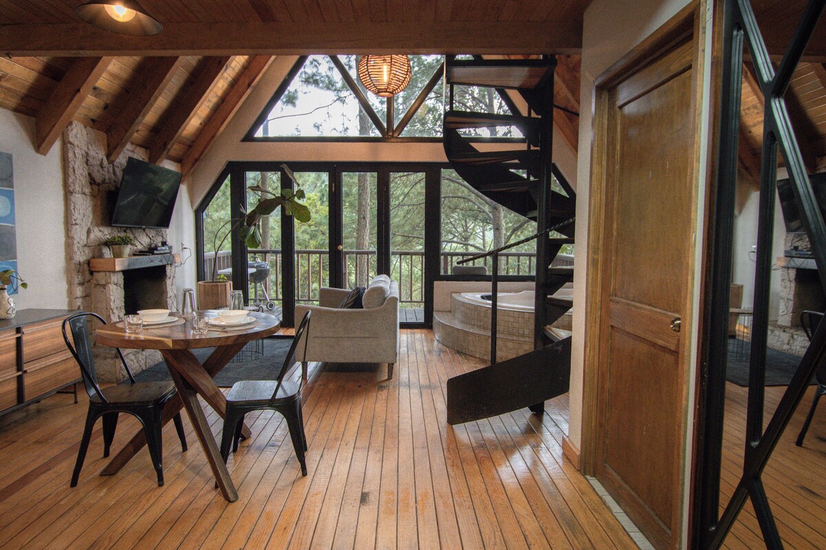 A spacious loft with a wooden floor, a dining area with a table and chairs, a sofa, a fireplace, and a large window overlooking a forest.