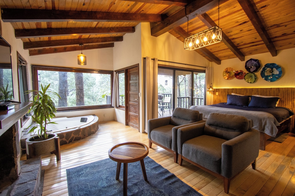 A spacious room with a wooden ceiling, a large bed, a seating area with two armchairs, a small round table, and a potted plant.