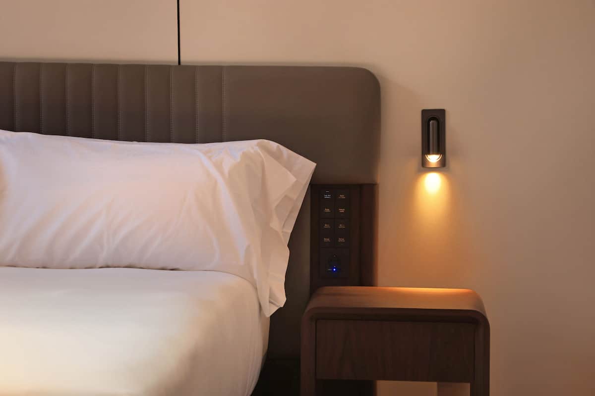 Una cama con ropa blanca, una mesita de noche con panel de control y una lámpara fijada a la pared.