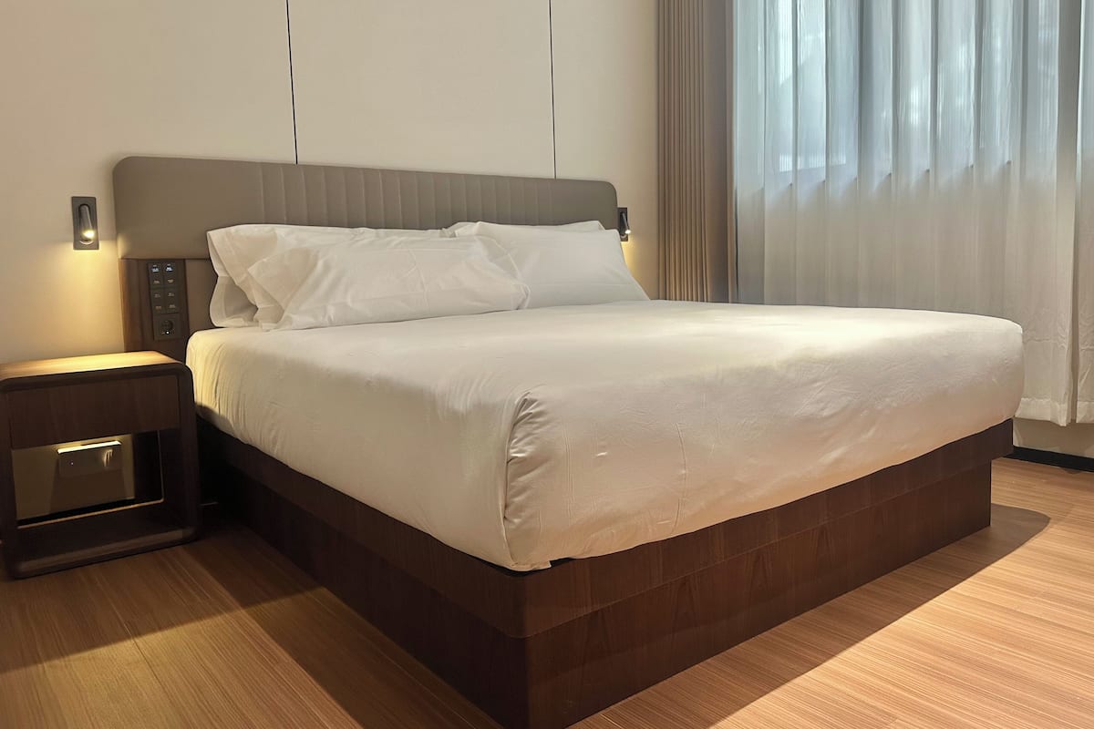 Una cama bien hecha con cabecera, una mesita de noche con lámpara y una ventana con cortinas.