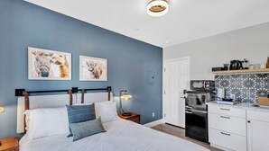 Ein Schlafzimmer mit einem Bett, Nachttischen und einem Schreibtischchen. Es gibt eine Küchenzeile mit einem Kühlschrank, einer Mikrowelle und einer Kaffeemaschine.