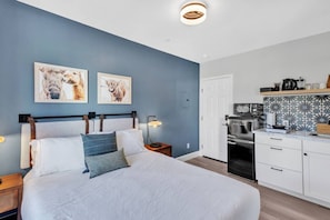 Ein Schlafzimmer mit einem Bett, Nachttischen und einem Schreibtischchen. Es gibt eine Küchenzeile mit einem Kühlschrank, einer Mikrowelle und einer Kaffeemaschine.