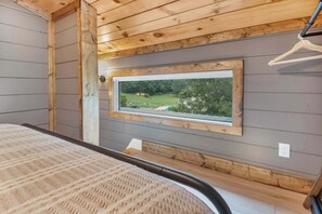 A bedroom with a wooden ceiling, a window with a view of a green landscape, a bed with a patterned cover, and a wooden floor. - 3 Cabins, Sleeps 18 - Private Hot Tubs, Near Chatt (Chattanooga)