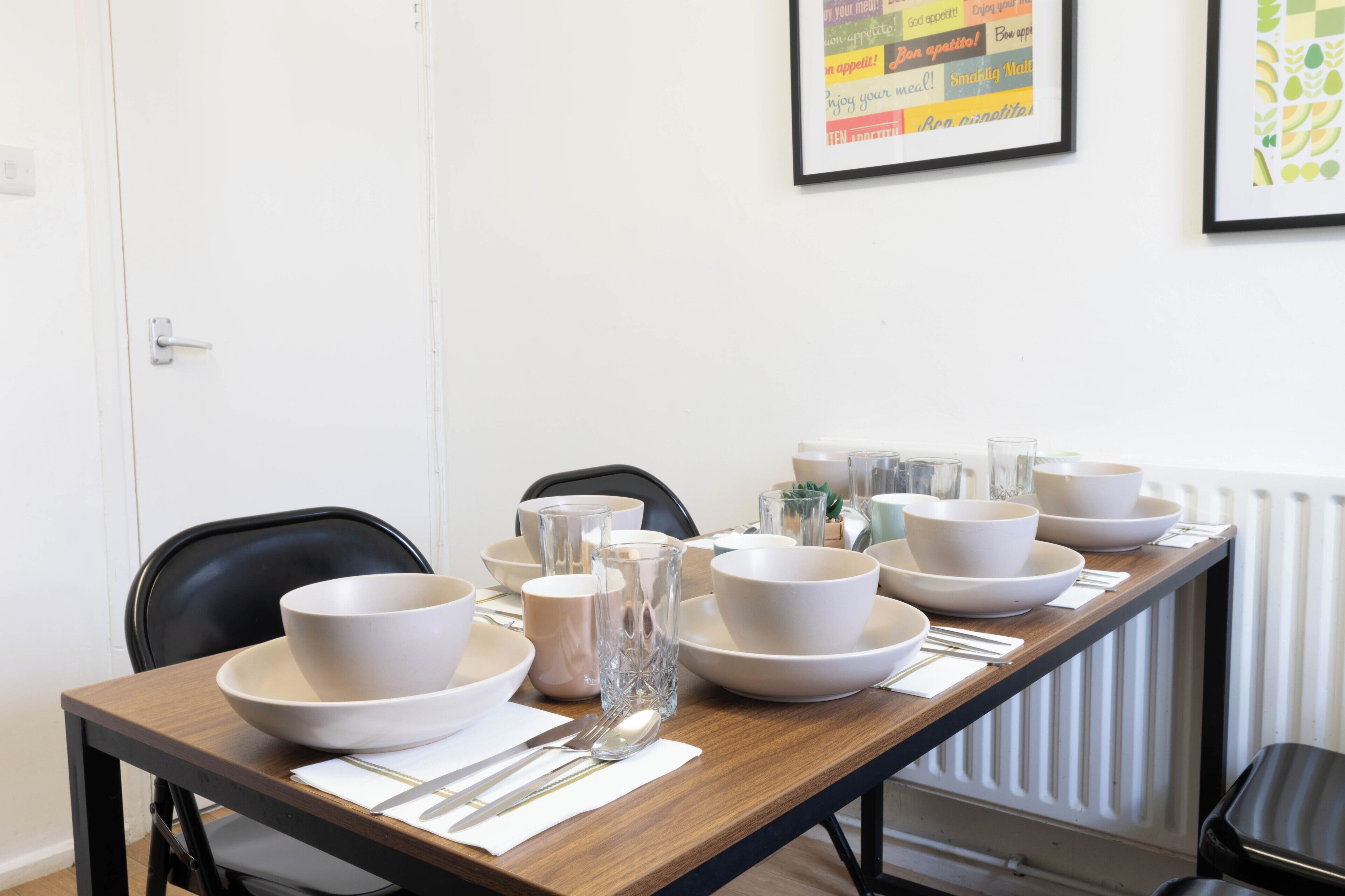 A dining table set with bowls, glasses, and cutlery, chairs, and framed artwork on the wall.