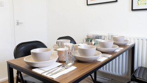 Ein Esszimmer mit einem gedeckten Tisch, Schüsseln, Gläsern, Besteck, Stühlen und einem gerahmten Kunstwerk an der Wand.