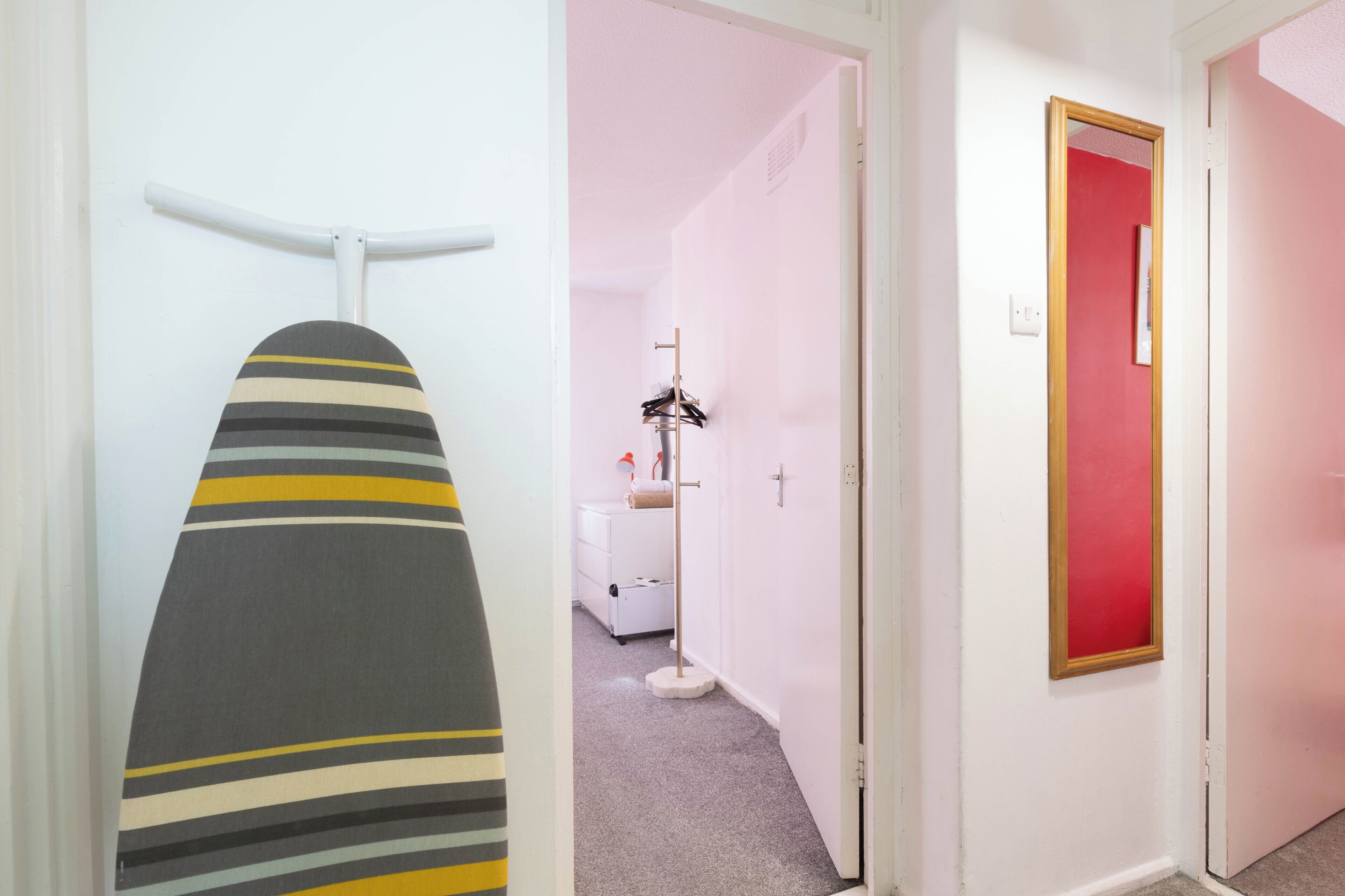 A hallway with a striped ironing board, a coat rack, and a red-framed mirror.