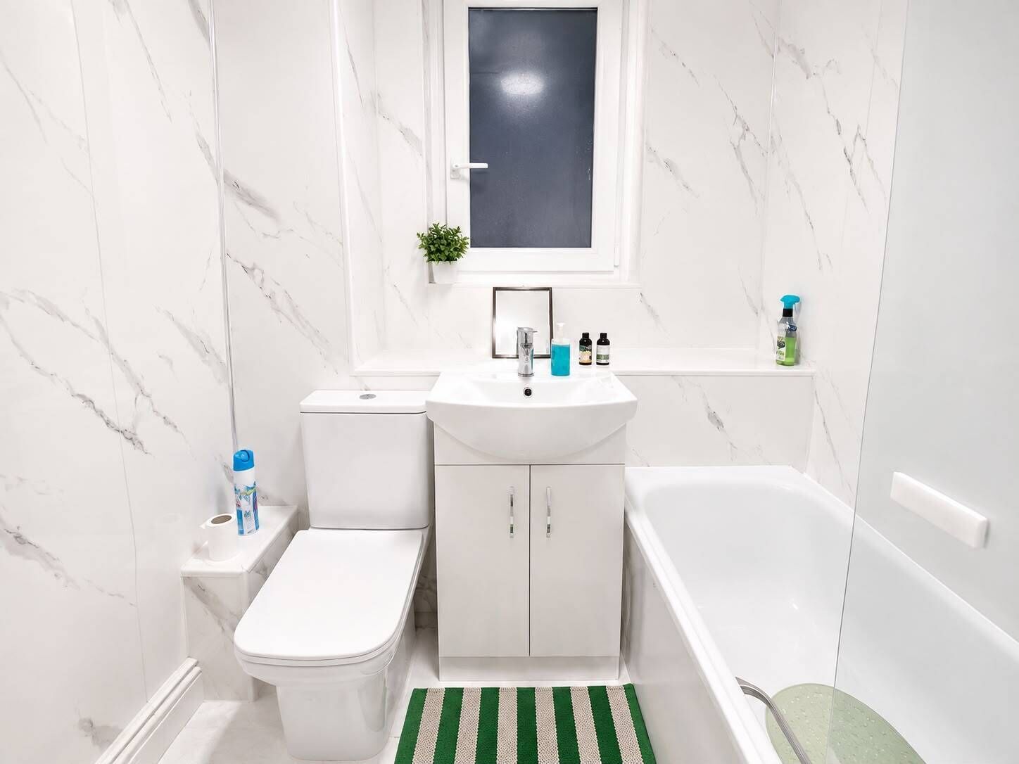 Baño con inodoro, lavamanos y bañera, paredes de mármol y un ventanal pequeño.