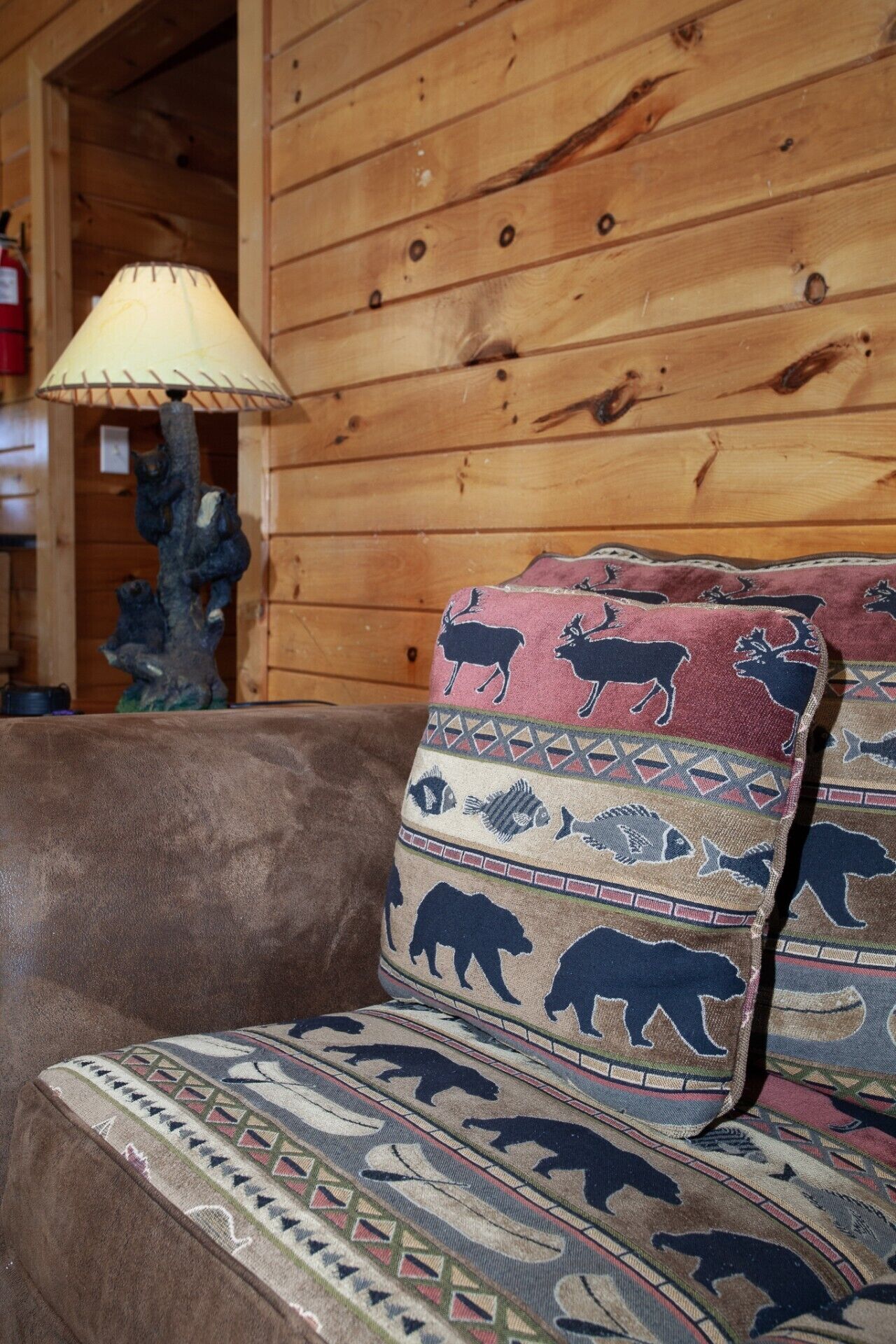 A wooden cabin interior with a brown sofa and patterned cushions, a lamp, and a decorative tree sculpture.