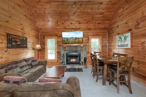 Ein Blockhaus-Innenraum mit Kamin, einem darüber montierten Fernseher, einer Couch mit gemusterten Kissen und einem Esstisch mit Stühlen.