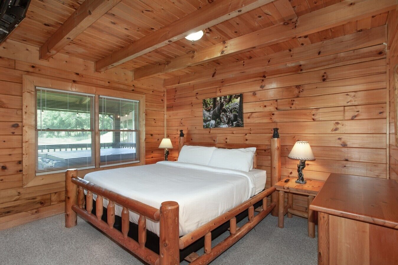 A wooden cabin bedroom with a bed, two bedside lamps, a window with blinds, and a picture on the wall.
