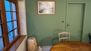 A room with a wooden table, a green wall, a framed picture, a door, and a window with a view. - Gîte de la Rivière, 3 stars, sleeps 2 to 6, Munster, Colmar, wine routes, and mountain ridges (Breitenbach-Haut-Rhin)