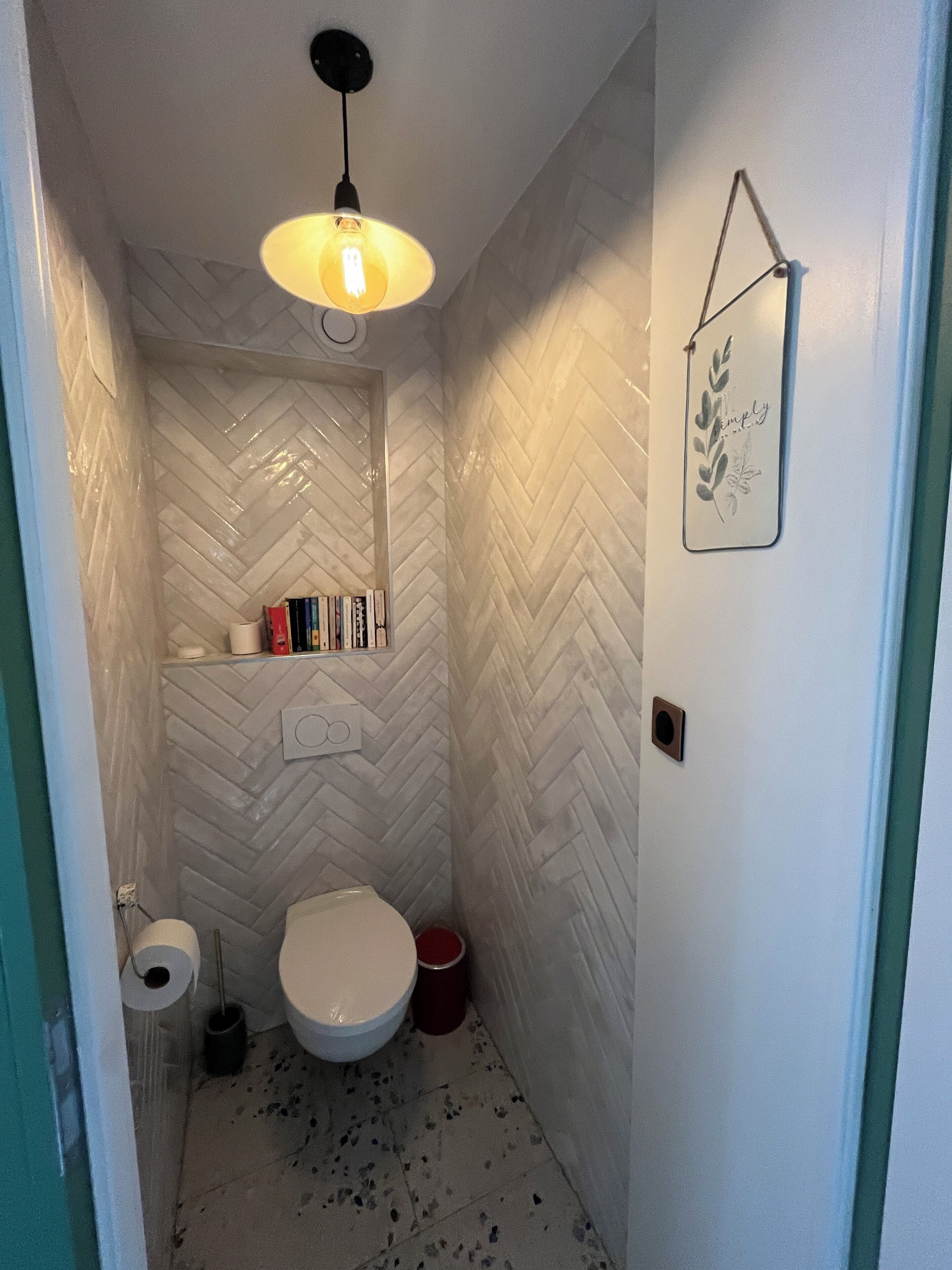 A small bathroom with a white herringbone tile wall, a toilet, a sink, and a hanging light fixture.