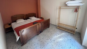 A bedroom with a wooden bed, a nightstand, a wall-mounted shelf with a white pillow, and a speckled tiled floor. - Gîte de la Rivière, 3 stars, sleeps 2 to 6, Munster, Colmar, wine routes, and mountain ridges (Breitenbach-Haut-Rhin)