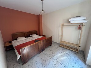 A bedroom with a wooden bed, a nightstand, a wall-mounted shelf with a white pillow, and a speckled tiled floor. - Gîte de la Rivière, 3 stars, sleeps 2 to 6, Munster, Colmar, wine routes, and mountain ridges (Breitenbach-Haut-Rhin)