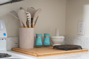 Una encimera de cocina con una taza blanca que contiene utensilios de madera, dos saleros y pimenteros color turquesa, un mortero y pilón blancos, y una sartén de hierro fundido negra sobre una tabla de madera.