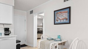 Una habitación compacta con kitchenette, un comedor con mesa y sillas, una cama al fondo y un cuadro en la pared.