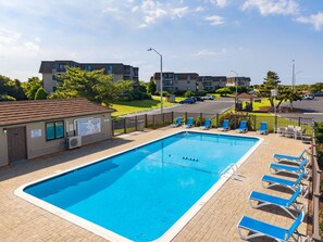 En rektangulär swimmingpool med en kant av kakel, omgiven av en terrass av tegelsten. Det finns solstolar och en liten lusthus.