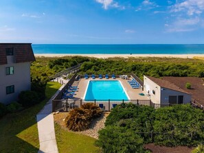 Une piscine avec vue sur la plage, entourée de verdure et bordée d’un bâtiment.
