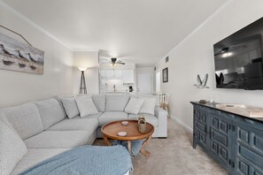 Ein modernes Wohnzimmer mit einem Ecksofa, einem runden Holztisch, einer Stehlampe und einem durch eine offene Tür einsehbaren Küchenbereich.
