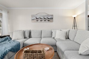 A living room with a grey sectional sofa, a blue throw blanket, a wicker basket on a wooden coffee table, and a floor lamp. - Southwinds Hideaway| Pool · Private Beach · 2BR AB (Atlantic Beach)