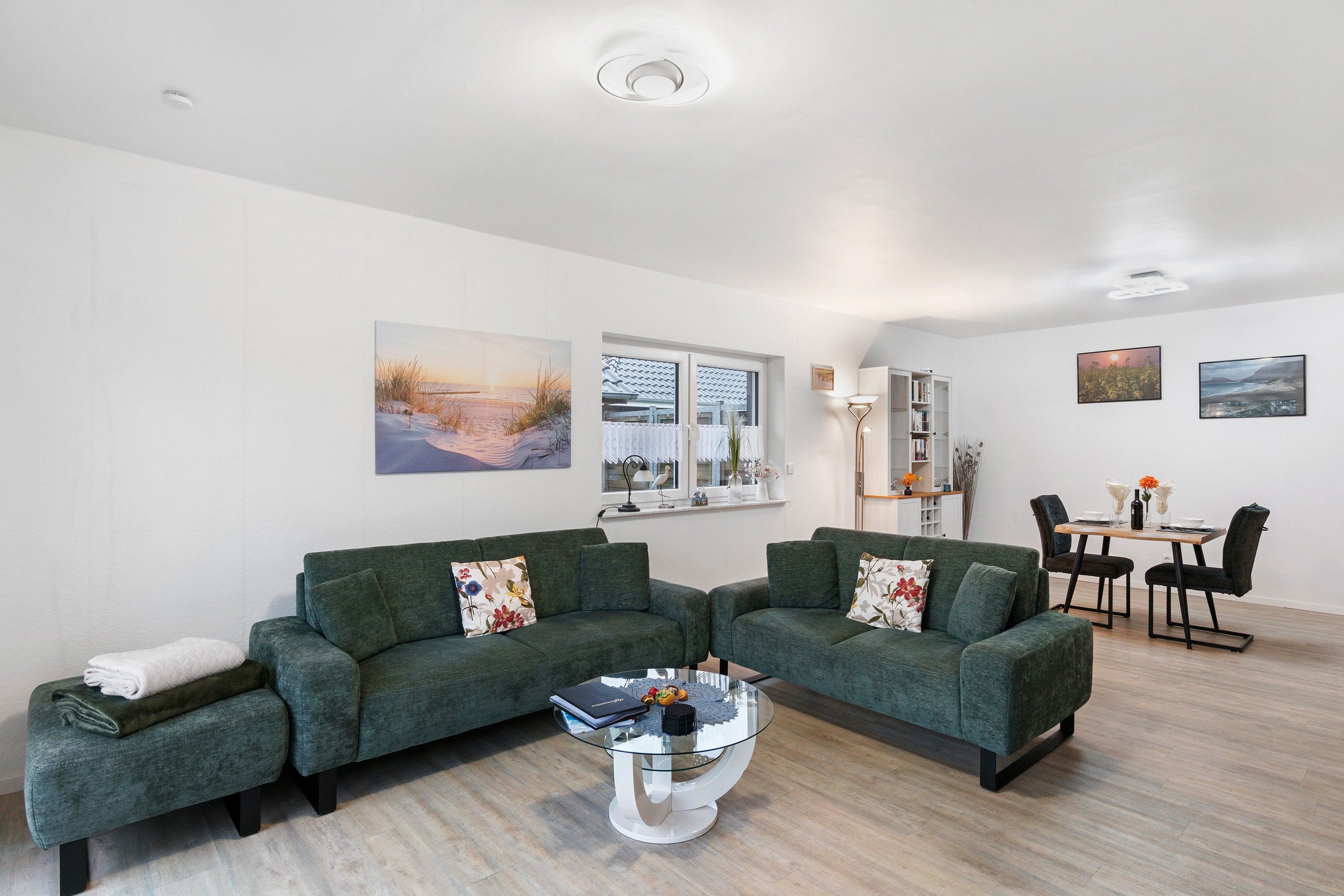 Ein modernes Wohnzimmer mit einer grünen Sofagarnitur, einem Glastisch und einem Essbereich mit weißem Tisch und Stühlen.