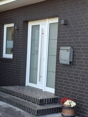 Ein Gebäude mit dunklen Ziegelsteinen, doppelverglasten Glastüren mit weißem Rahmen und einem kleinen Briefkasten an der Wand.