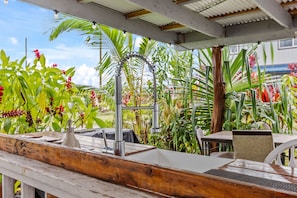 A wooden deck with a metal sink, surrounded by lush tropical plants and a covered area with chairs and a table.