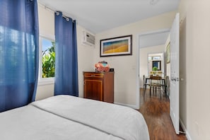 Une chambre à coucher avec un lit, une commode en bois, un coin repas avec des chaises et un tableau au mur.