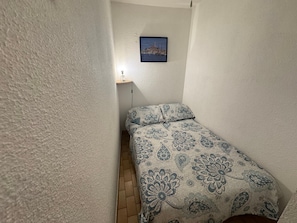 Ein kleiner, schmales Zimmer mit einem Bett, einem Wandbild und einem kleinen Tisch mit einer Lampe.