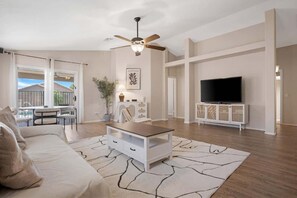 Un salon comprenant un canapĂ©, une table basse, un tĂ©lĂ©viseur, un ventilateur de plafond et une vue sur lâextĂ©rieur.