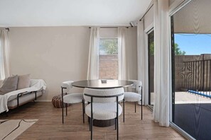Une piĂšce comprenant une table ronde, des chaises Ă coussins blancs, un canapĂ© et une porte-fenĂȘtre coulissante donnant sur un espace extĂ©rieur.