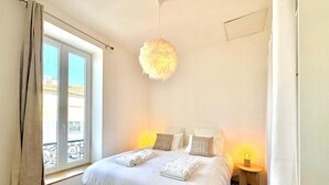 Un dormitorio con una cama, dos mesitas de noche, una lámpara colgante y un ventanal con cortinas.