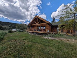 A wooden cabin with a balcony, surrounded by a grassy field and trees. - River Valley Lodge - 79393 Brae Gate (Portola)