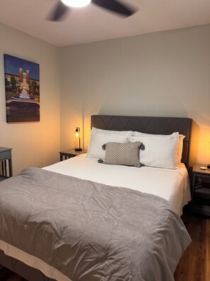 Una habitación de hotel con una cama grande, un cabecero, una mesita de noche, una lámpara y un cuadro de un campus universitario.