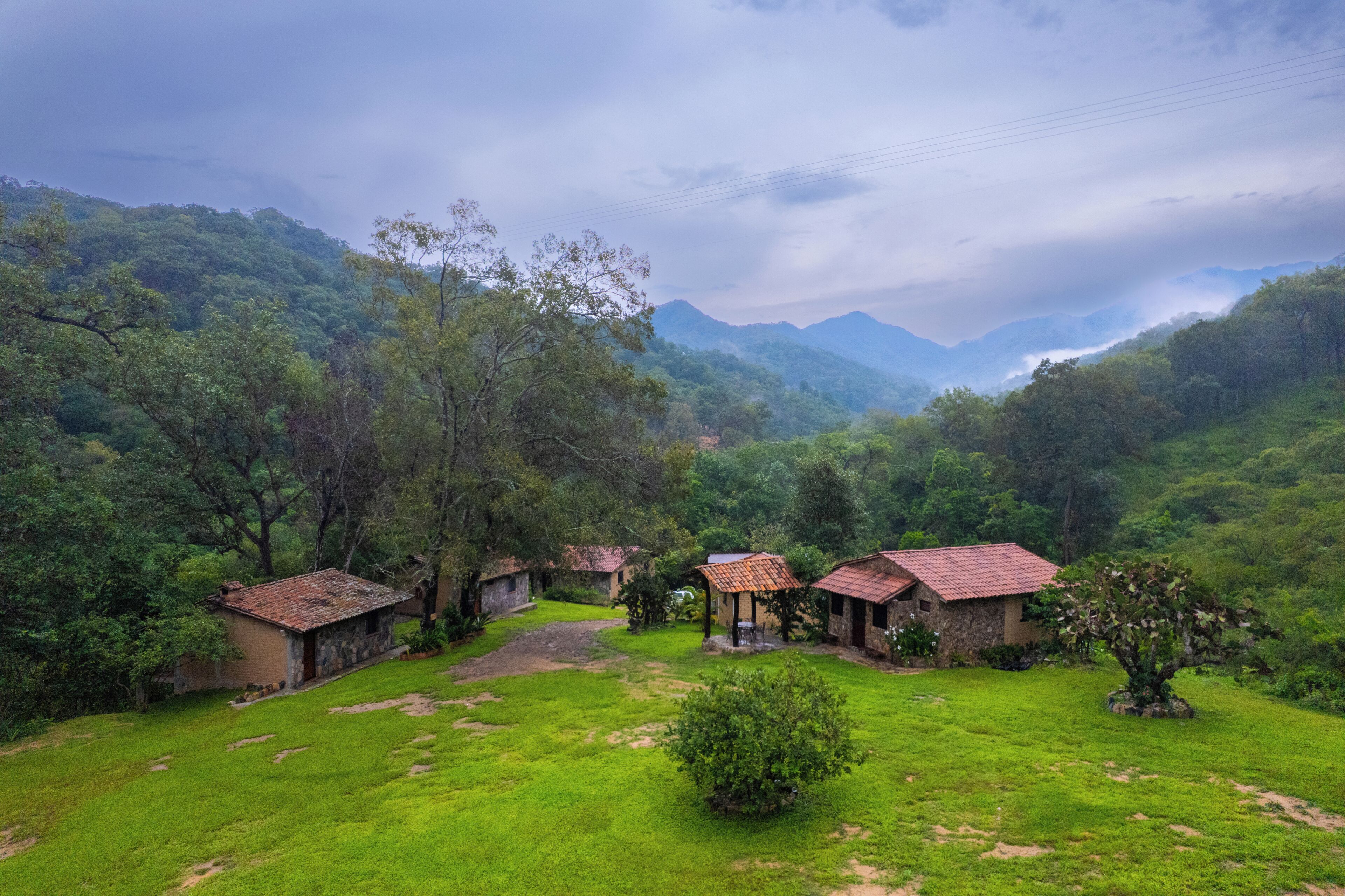 Paisagem rural com vários edifícios rústicos rodeados por vegetação exuberante e montanhas ao fundo.