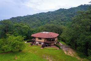 Uma casa grande de dois andares com telhado de telhas vermelhas, rodeada por vegetação exuberante e colinas.