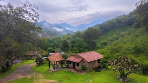 Paisaje rural con una casa de piedra y una construcción más pequeña con techo rojo, rodeado de vegetación exuberante y montañas al fondo.