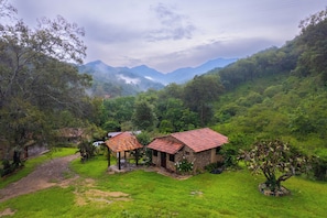 Paisaje rural con una casa de piedra y una construcción más pequeña con techo rojo, rodeado de vegetación exuberante y montañas al fondo.