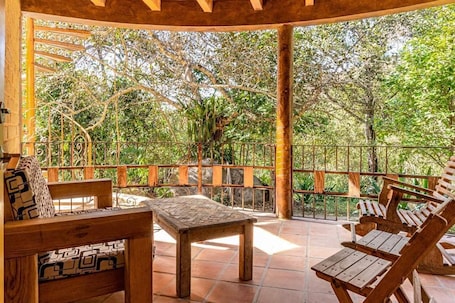 A covered outdoor seating area with wooden furniture and a view of trees.