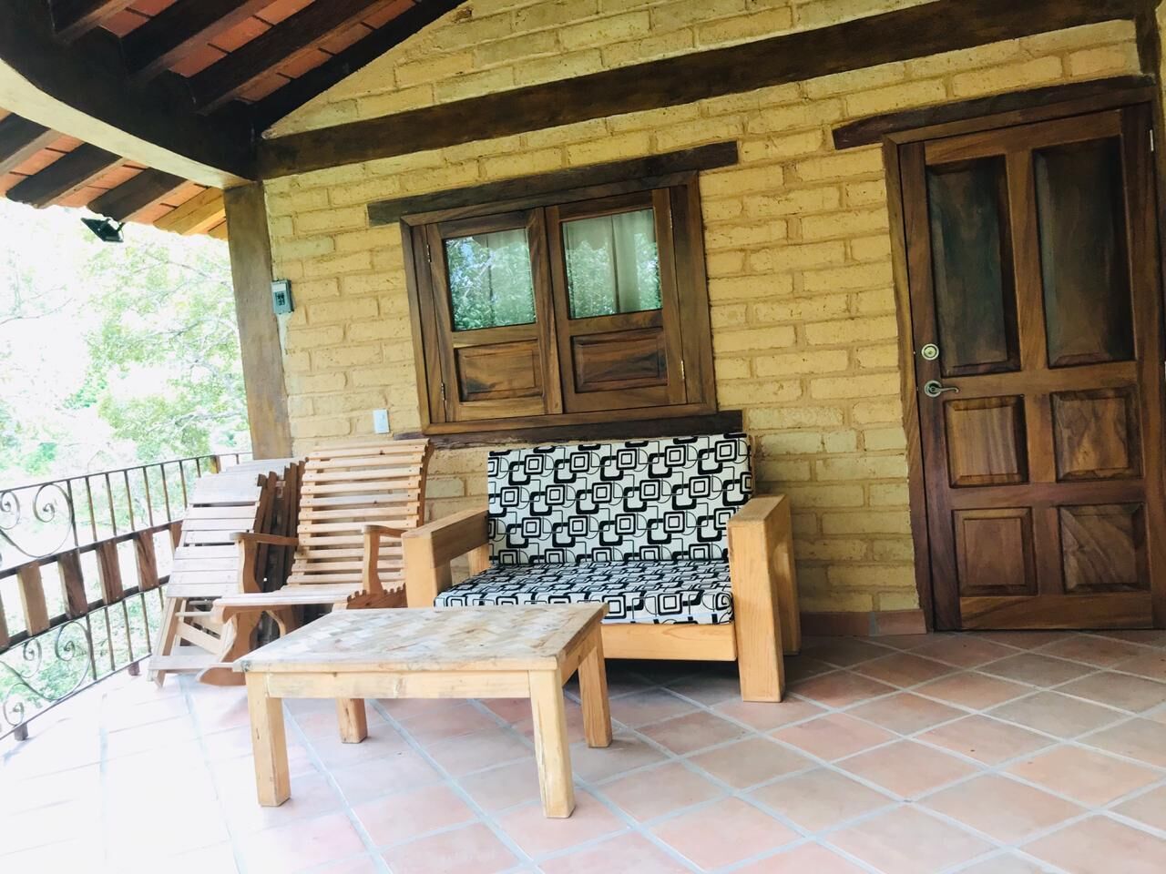 A covered patio with wooden furniture, a brick wall, and a wooden door.