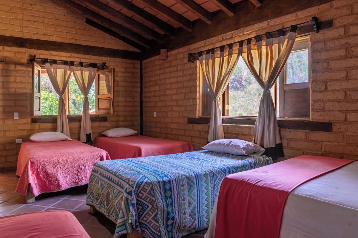 Une chambre avec quatre lits, chacun recouvert d’une couvre-lit à motifs différents, des poutres en bois au plafond et des fenêtres avec des rideaux.
