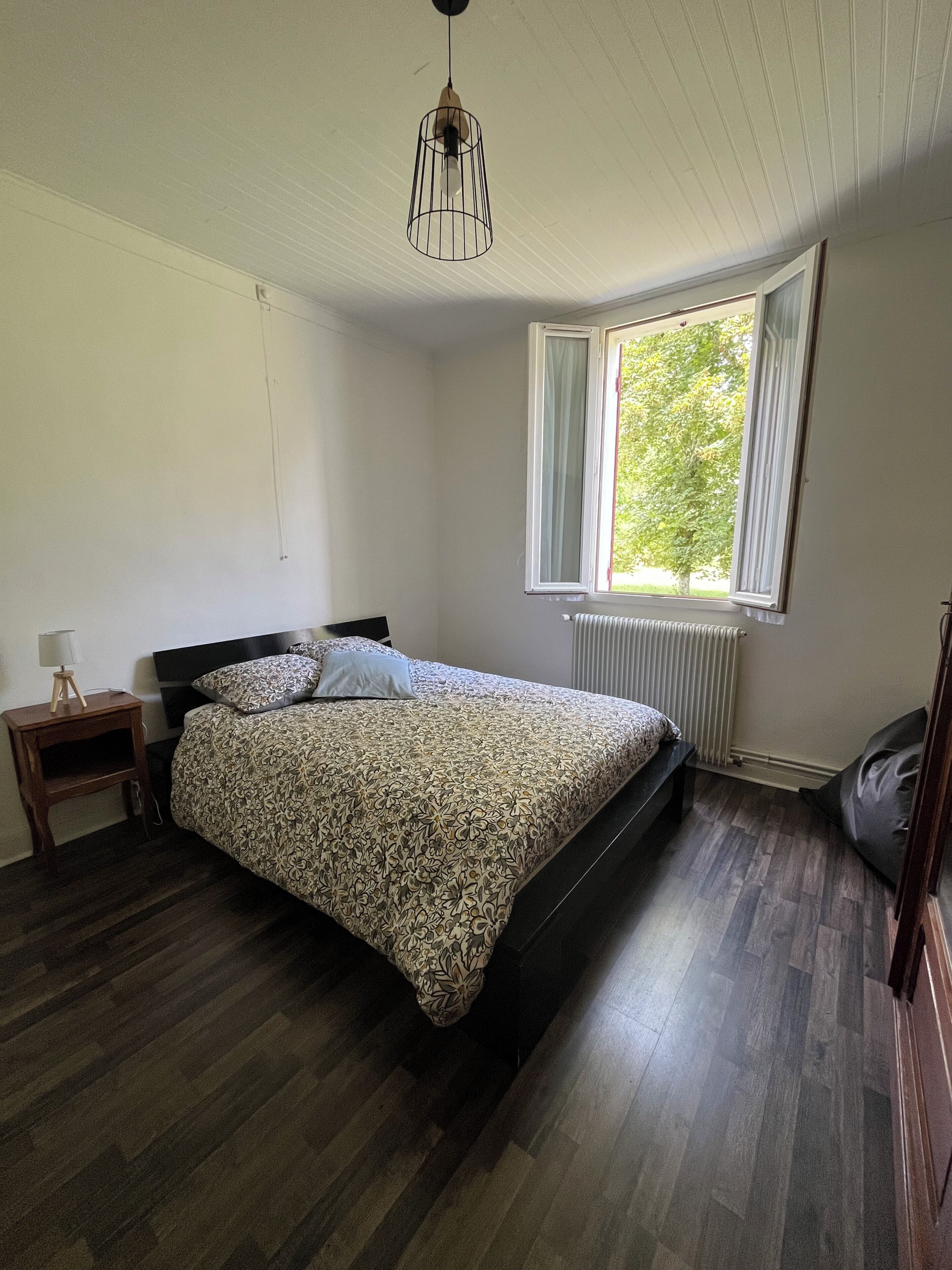 Ein Schlafzimmer mit Bett, Nachttisch, Heizkörper, Fenster und einer Pendelleuchte.